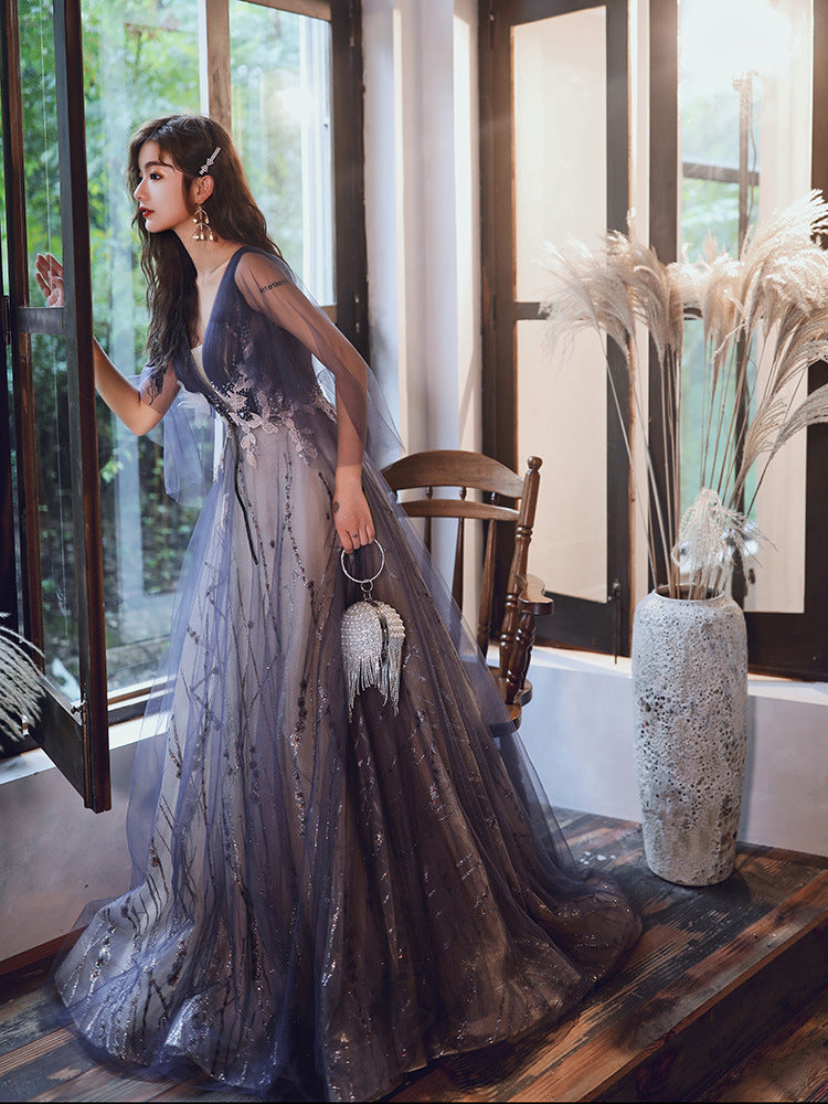 Woman in a long, elegant dress standing indoors with large windows and decorative elements.