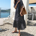 Woman in a black and white patterned dress holding a brown bag on a pebbled surface with a scenic background.