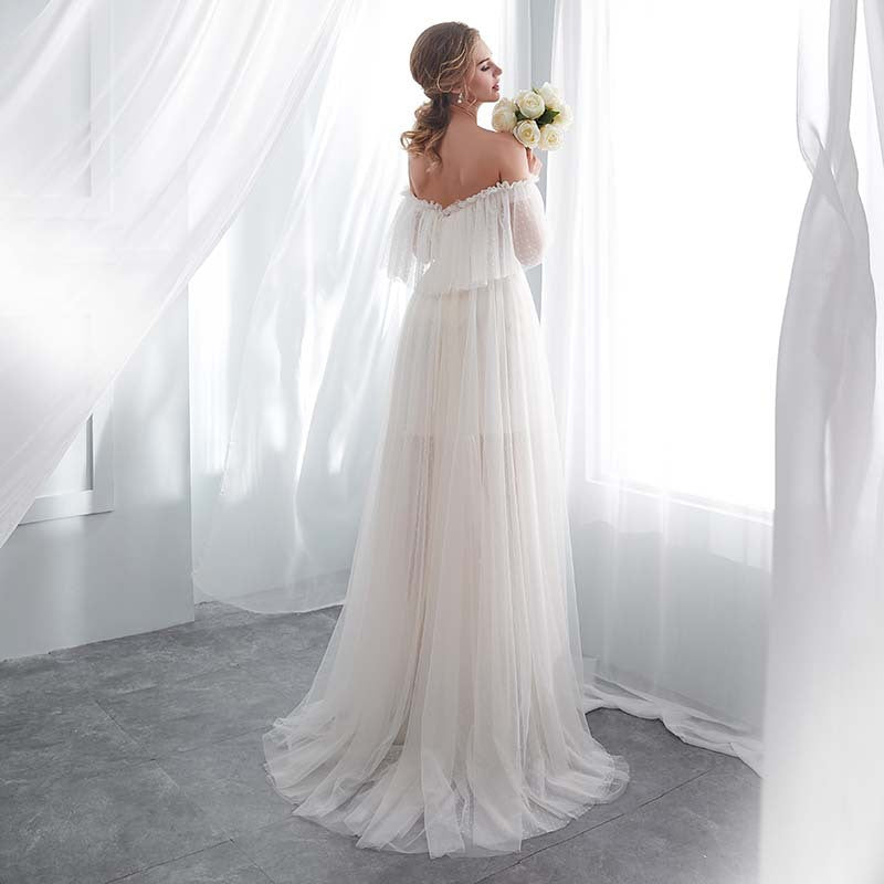 Woman in a white wedding dress holding flowers in a bright room with sheer curtains.