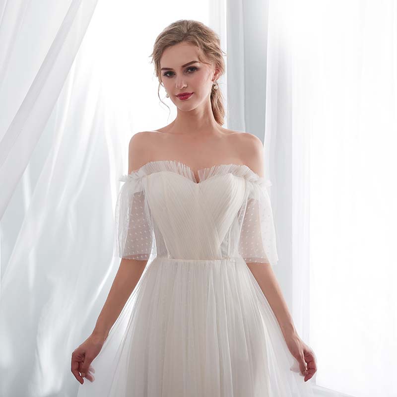Woman wearing a white off-shoulder dress with sheer sleeves in front of white curtains.