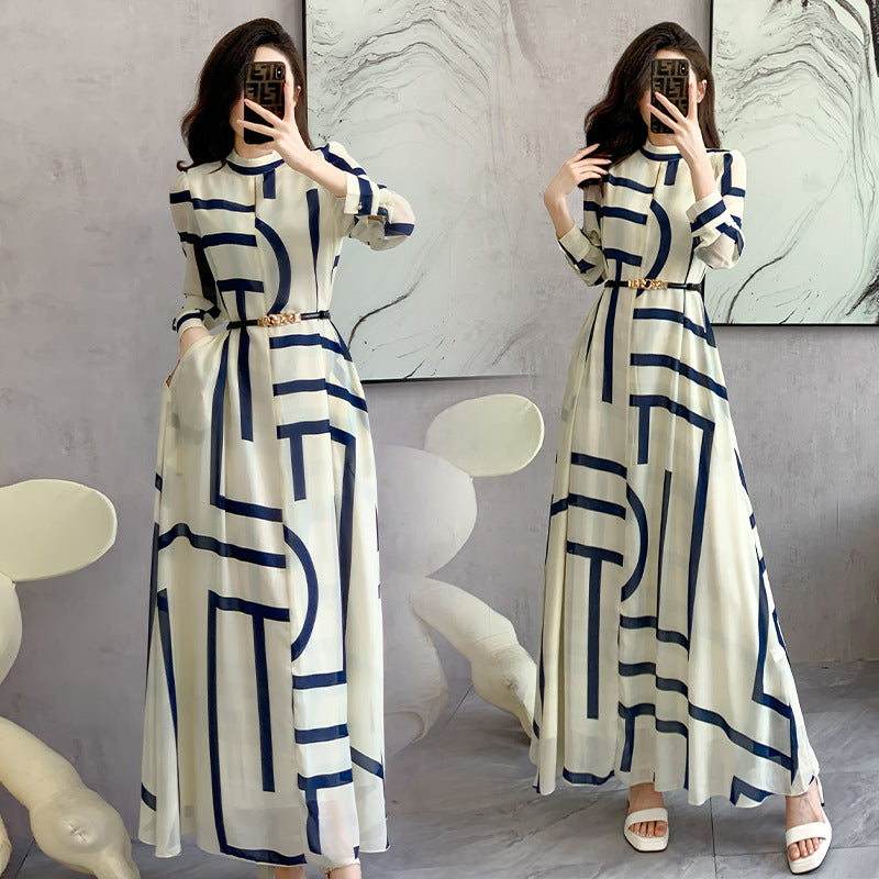 Woman wearing a long white dress with blue geometric patterns, standing in front of a marble wall.