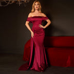 Woman in a burgundy off-shoulder gown posing against a dark background with a red couch.