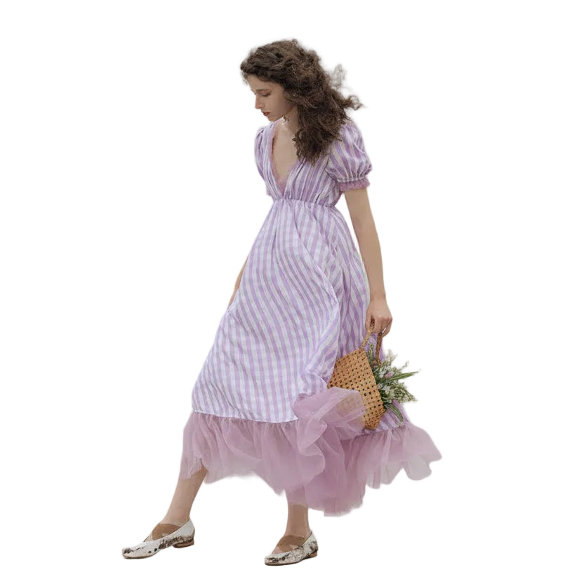 Woman in a pink and white checkered dress walking near a body of water with mountains in the background.