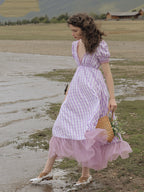 Woman in a pink and white checkered dress walking near a body of water with mountains in the background.