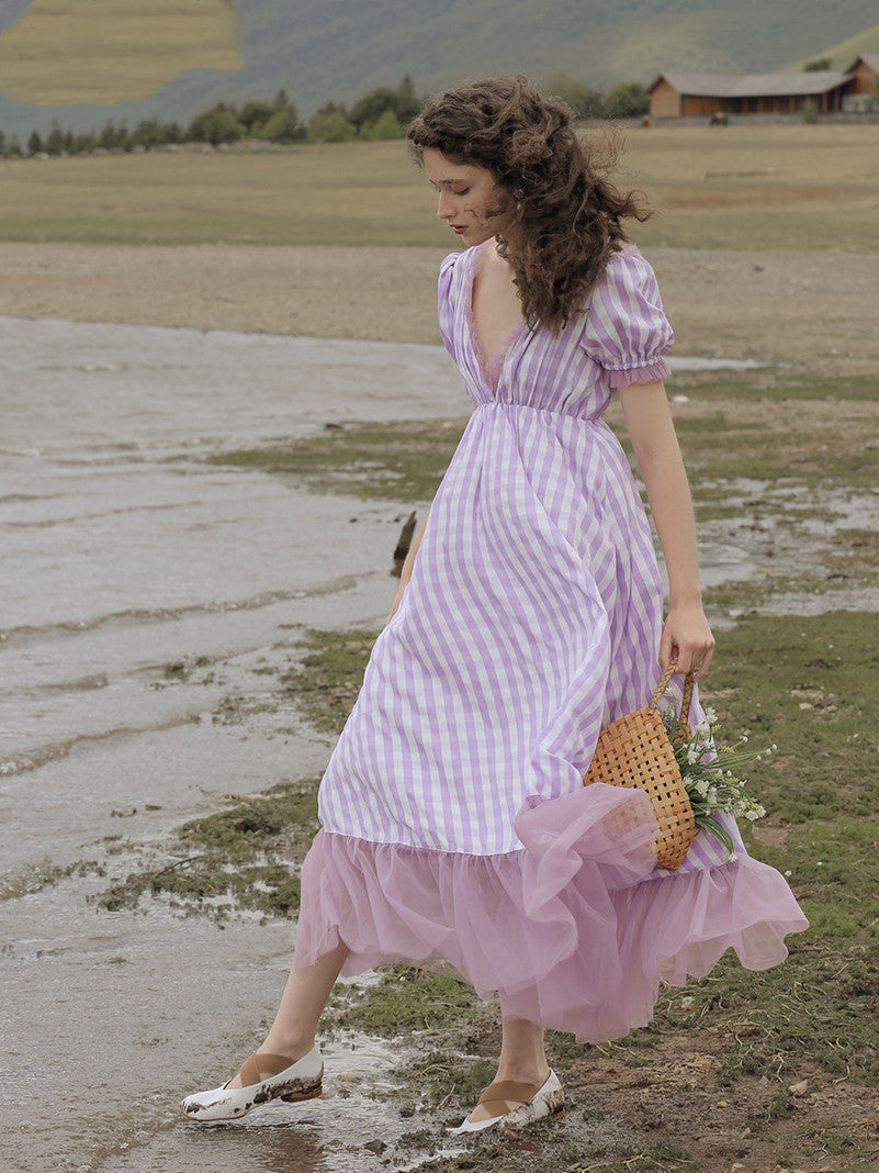 Woman in a pink and white checkered dress walking near a body of water with mountains in the background.